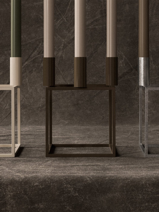 A close-up of ivory, brown, and silver Kubus 4 candle holders with olive and beige candles on a dark, textured surface.