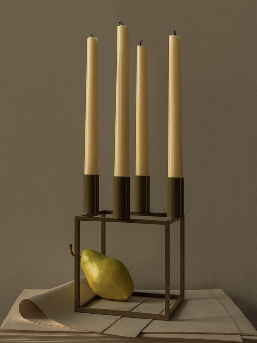 The olive-coloured Kubus 4 candle holder holds four tall cream candles. A green pear is resting inside its geometric base.