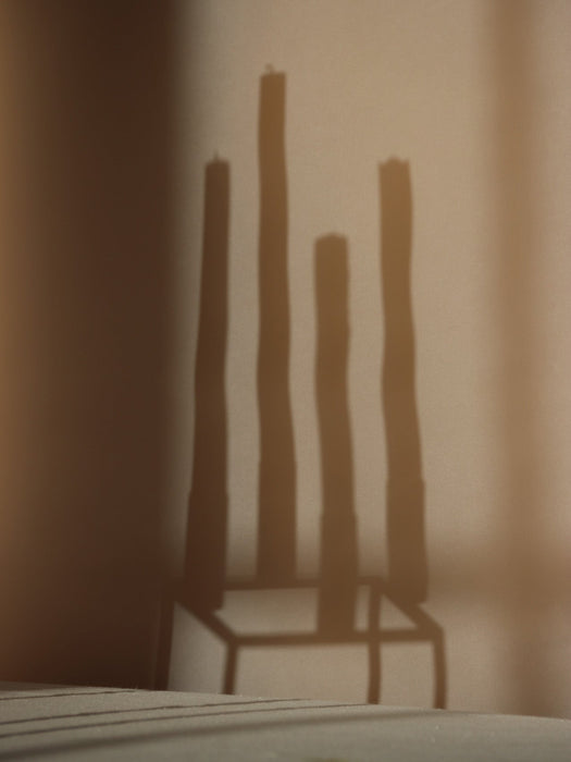 Atmospheric close-up showing the distinct shadow of the Kubus 4 candleholder and four tall candles cast on a warm-toned wall.