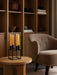 The black Kubus 4 candle holder with lit candles on a wooden table, next to a beige Ingeborg Lounge Chair in a cosy room.
