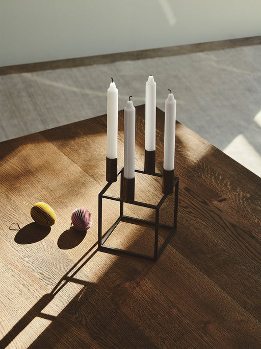 Black by Lassen Kubus 4 candle holder with four white candles on a sunlit dark wood table next to two paper ornaments.