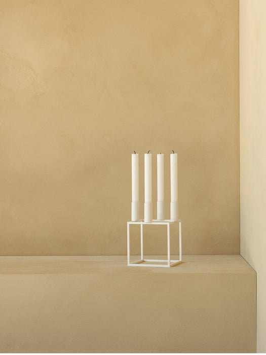 A white, geometric Kubus Candle Holder holding four tall white candles on a beige surface against a matching beige wall.