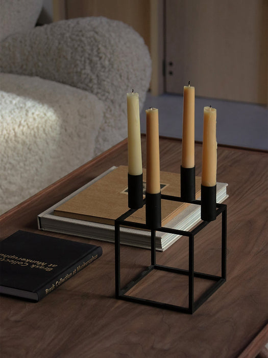 A black Kubus 4 candle holder with four beige candles, styled on books on a wooden Jager Lounge Table with a Tired Man chair.