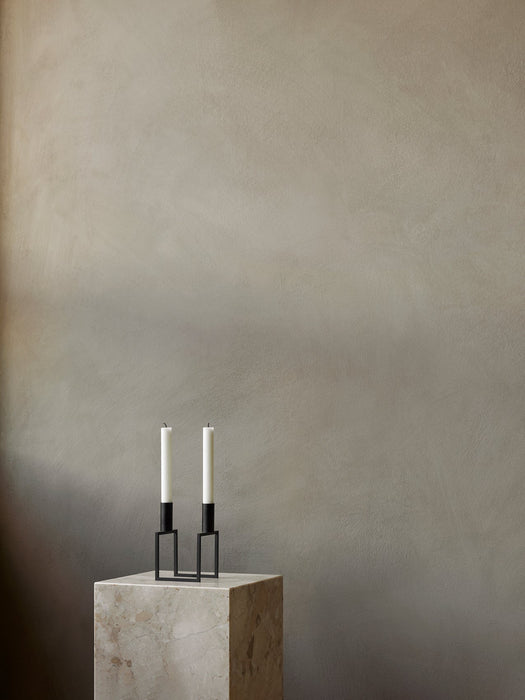 A black Kubus Line Candle Holder with two white candles on a marble Plinth Pedestal against a muted, textured beige wall.