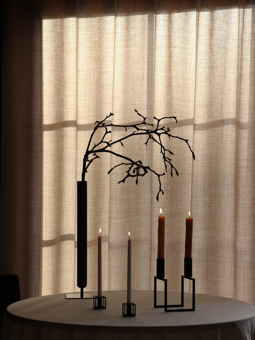 A still life with a Stance Vase holding a branch and lit candles in Kubus Line and Kubus Mikro holders on a softly lit table.