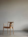 A Merkur Dining Chair with Armrests in natural oak with a tan leather seat, standing in a minimalist room with a grey wall.