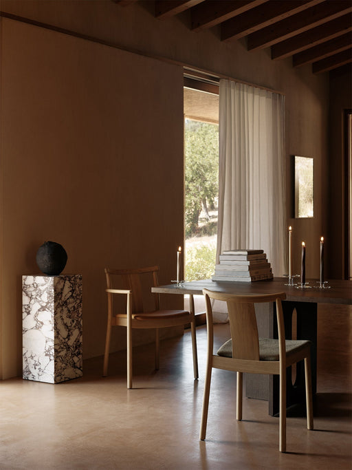 Two light oak Merkur Dining Chairs at a dark Androgyne Dining Table in a warmly lit room with a marble Plinth Pedestal.