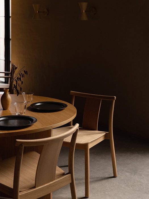 A warmly lit dining room with two oak Merkur Dining Chairs at a round Androgyne Table, with Collector Wall Lamps on the wall.