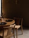 A warmly lit dining room with two oak Merkur Dining Chairs at a round Androgyne Table, with Collector Wall Lamps on the wall.