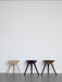 Three sculptural ML 42 Stools in light oak, black, and dark brown wood finishes, displayed in a row against a white wall.