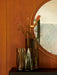Nimbus Mirror, a round brass-framed mirror on a wood wall, styled with two dark glass vases holding flowers.