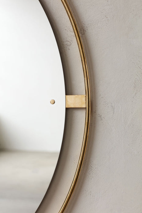 A close-up of the Nimbus Mirror, highlighting the polished brass halo frame and mounting detail against a textured grey wall.