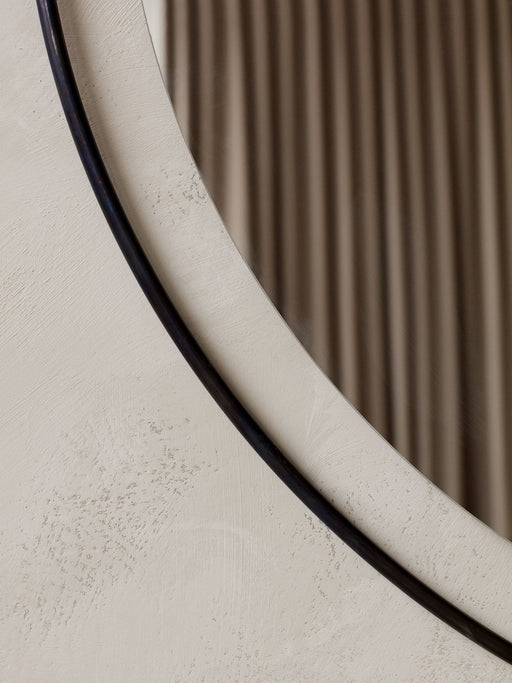 A close-up of the Nimbus Mirror, showing its slim, dark frame against a textured off-white wall, reflecting a soft curtain.