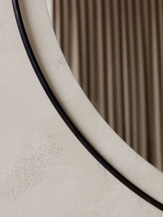 A close-up of the Nimbus Mirror, showing its slim, dark frame against a textured off-white wall, reflecting a soft curtain.