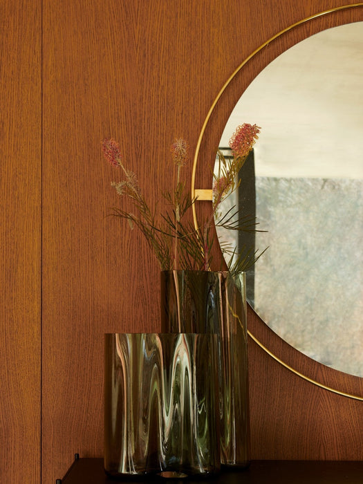 A close-up view of the round, brass-framed Nimbus Mirror hanging on a dark oak wall, with two smoky glass Aer Vases in front.