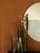 A close-up view of the round, brass-framed Nimbus Mirror hanging on a dark oak wall, with two smoky glass Aer Vases in front.