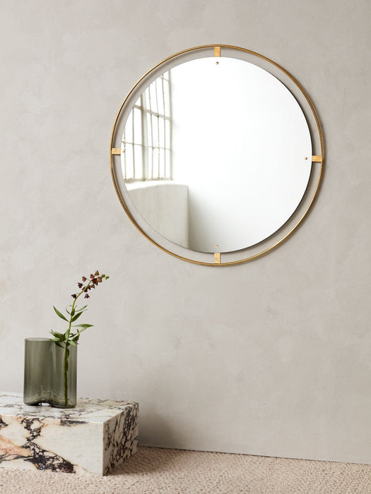 The round Nimbus Mirror in a brass frame hangs on a grey wall above a marble Plinth holding a grey Aer Vase with a flower.