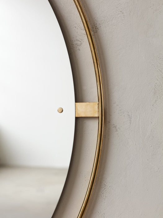 Close-up of the round Nimbus Mirror, highlighting the polished brass frame and mounting bracket against a light grey wall.