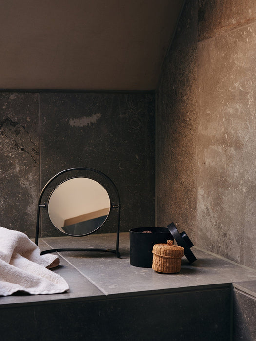 A black-framed Nimbus Table Mirror sits on a dark stone countertop in a bathroom, with a towel and storage baskets nearby.