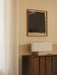 Nonna Table Lamp, travertine base and rectangular shade, sits on a dark wood sideboard beneath an abstract painting.