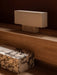 The Nonna Table Lamp in travertine with its rectangular sand-coloured shade, on a wooden shelf with a marble block below.