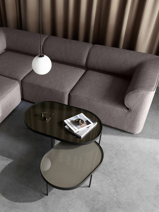 A modern living room with two glass NoNo Tables nested by a grey sofa, with a hanging lamp and taupe curtain.