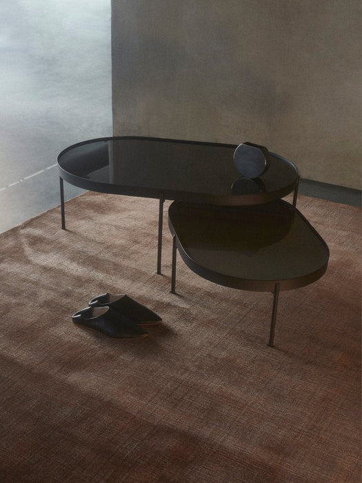 Two oval NoNo Tables, one large and one small, with dark glass tops, styled in a room on a brown rug with a Moon Vase.