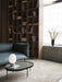 The NoNo Table with a glass top and JWDA lamp sits on a beige rug next to a teal sofa, in front of a large wooden bookcase.
