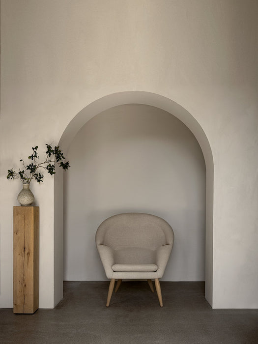 The Oda Lounge Chair, upholstered in a light fabric with wooden legs, sits inside an arched alcove next to a wooden plinth.