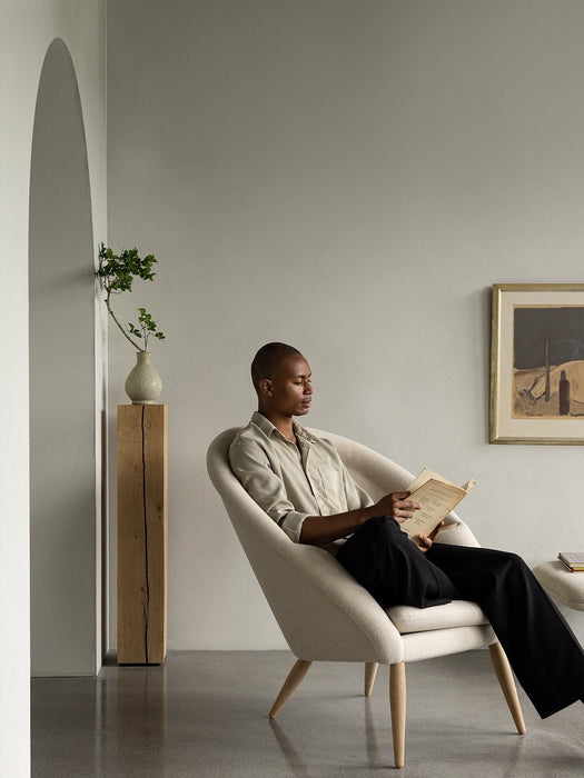 A man reads a book in the off-white Oda Lounge Chair. The room has grey walls, a wooden plinth, and an arched doorway.