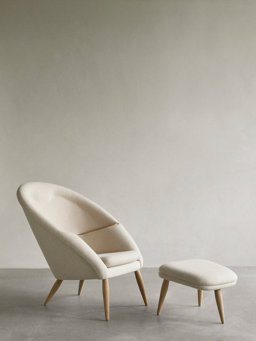 The Oda Lounge Chair and matching Oda Ottoman, upholstered in a cream fabric with natural oak legs, in a minimalist room.