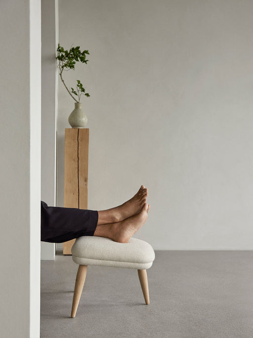A person rests their bare feet on the Oda Ottoman with its light fabric upholstery and natural oak legs in a minimal setting.