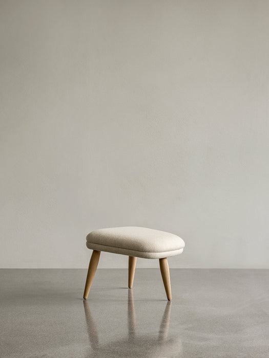 A frontal view of the Oda Ottoman with a cream textured cushion and splayed natural oak legs on a reflective grey floor.