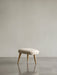 A frontal view of the Oda Ottoman with a cream textured cushion and splayed natural oak legs on a reflective grey floor.