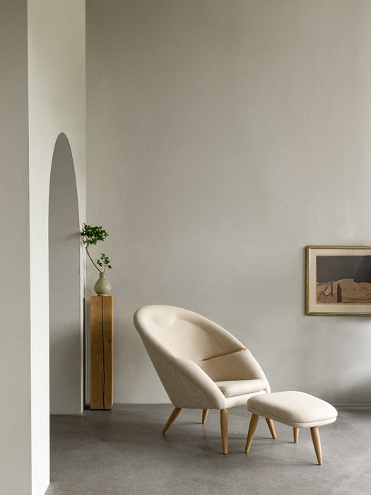 The Oda Lounge Chair and Oda Ottoman in cream fabric, styled in a minimalist room with grey walls and an arched doorway.