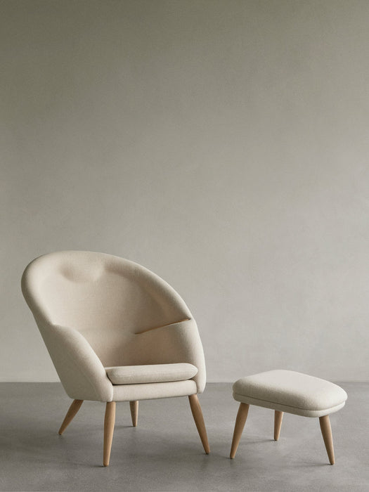 A cream-coloured Oda Lounge Chair with a curved back and oak legs, sits next to the matching Oda Ottoman on a grey floor.