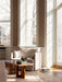 A beige armchair and a solid wood coffee table in a sun-drenched room with large windows and a herringbone parquet floor.