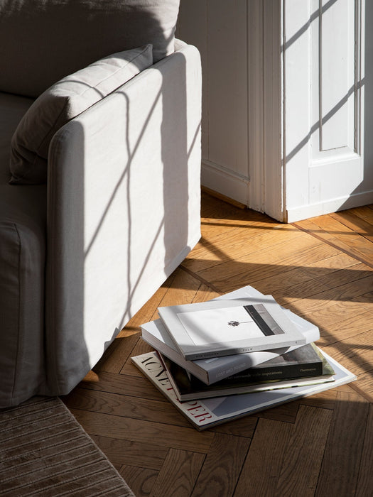 Sunlight casts a window shadow on the arm of the Offset Sofa w. Loose Cover. A pile of books sits on the herringbone floor.