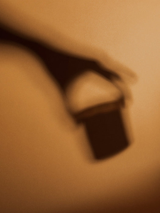 An artistic, blurry shadow of a hand holding the Olfacte Scented Candle, Chapter, against a warm, textured background.