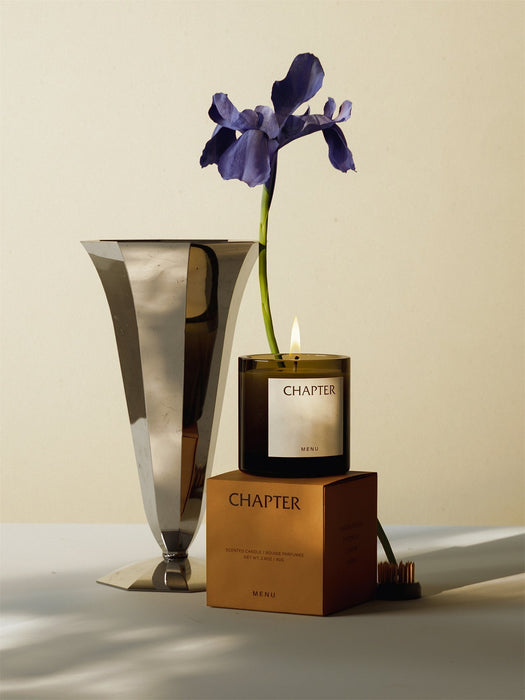 A lit MENU Olfacte Scented Candle, Chapter, sits on its box next to a metallic vase holding a single purple iris flower.