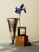 A lit MENU Olfacte Scented Candle, Chapter, sits on its box next to a metallic vase holding a single purple iris flower.