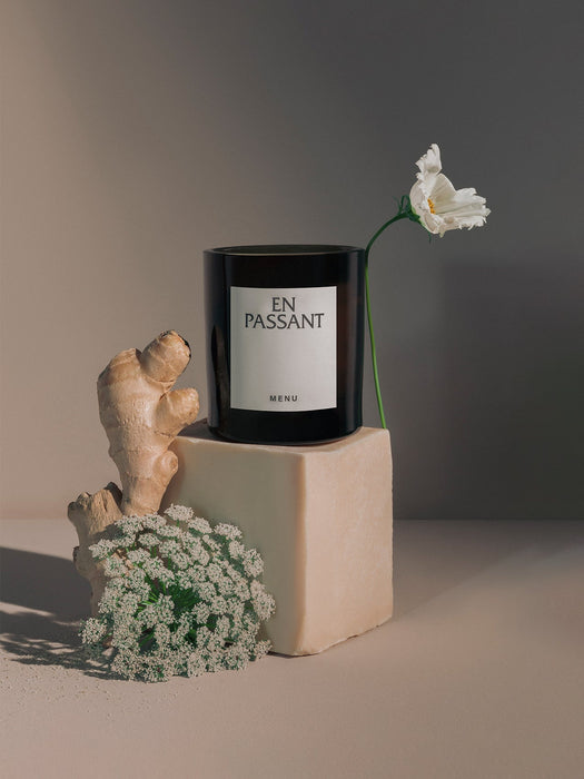 The Olfacte Scented Candle, En Passant, in a black jar, styled on a beige plinth with ginger root and delicate white flowers.