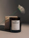 The MENU Olfacte Scented Candle, En Passant, in a dark glass jar, shot with a blurred white flower in the foreground.