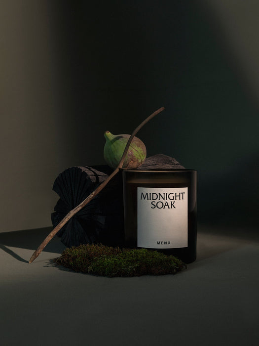 The MENU Olfacte Scented Candle in Midnight Soak, styled in a moody setting with charcoal, a green fig, moss, and a twig.