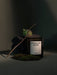 The MENU Olfacte Scented Candle in Midnight Soak, styled in a moody setting with charcoal, a green fig, moss, and a twig.