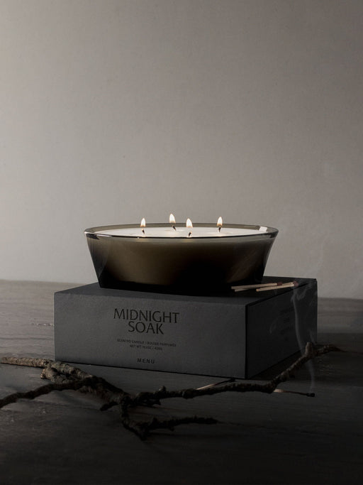 The four-wick Olfacte Scented Candle, Midnight Soak, glows in a dark glass bowl atop its black packaging. A twig rests below.