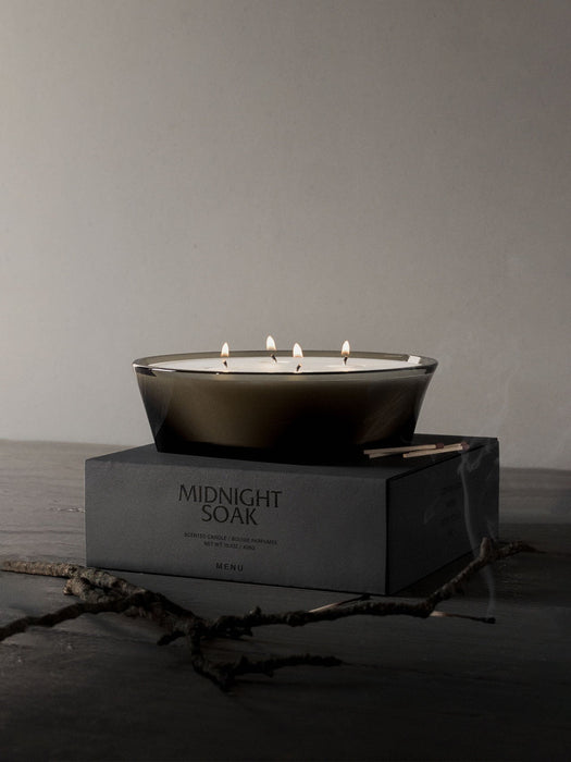 The four-wick Olfacte Scented Candle, Midnight Soak, glows in a dark glass bowl atop its black packaging. A twig rests below.