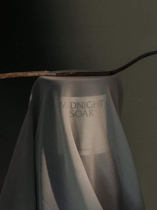 An artistic photograph of the Olfacte Scented Candle, Midnight Soak, shrouded in a light grey fabric with its label visible.