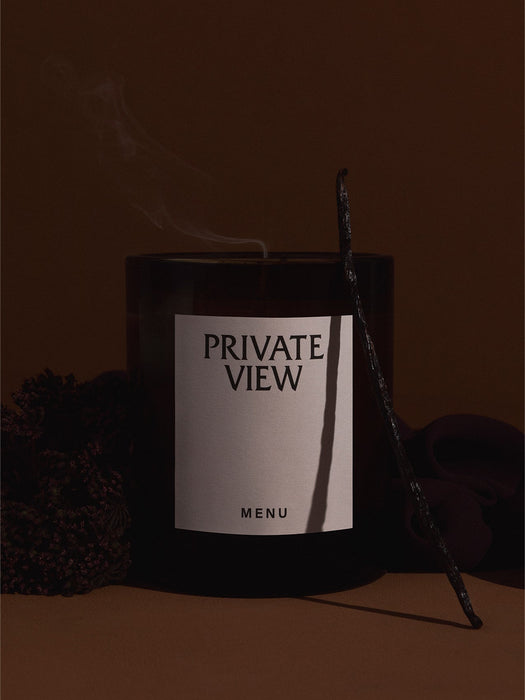 Olfacte 'Private View' scented candle in a dark glass jar with smoke rising. Styled with a vanilla pod and dried flowers.