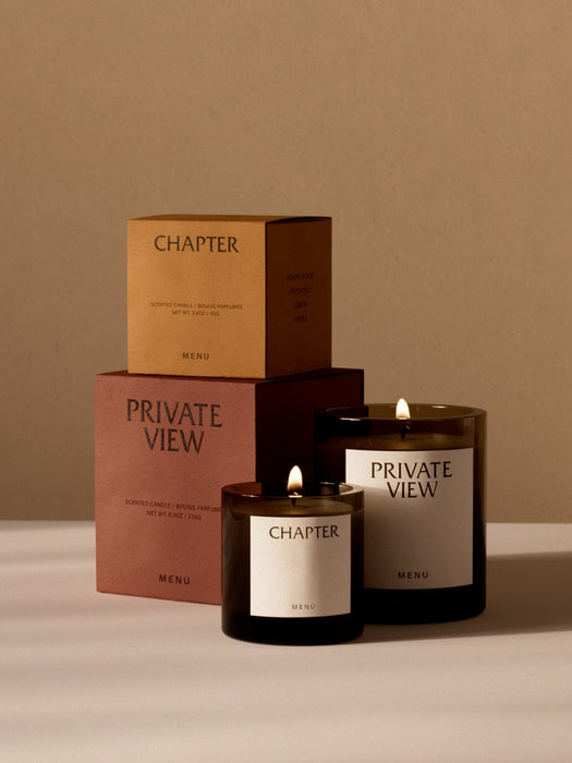 Two lit Olfacte scented candles, Chapter and Private View by Audo, in dark glass jars with their tan and terracotta boxes.